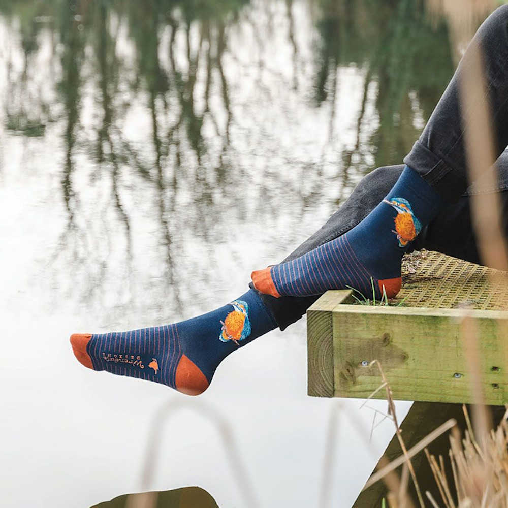 Person mit blauen Motivsocken und Sportschuhen sitzt entspannt auf einem Holzsteg am See